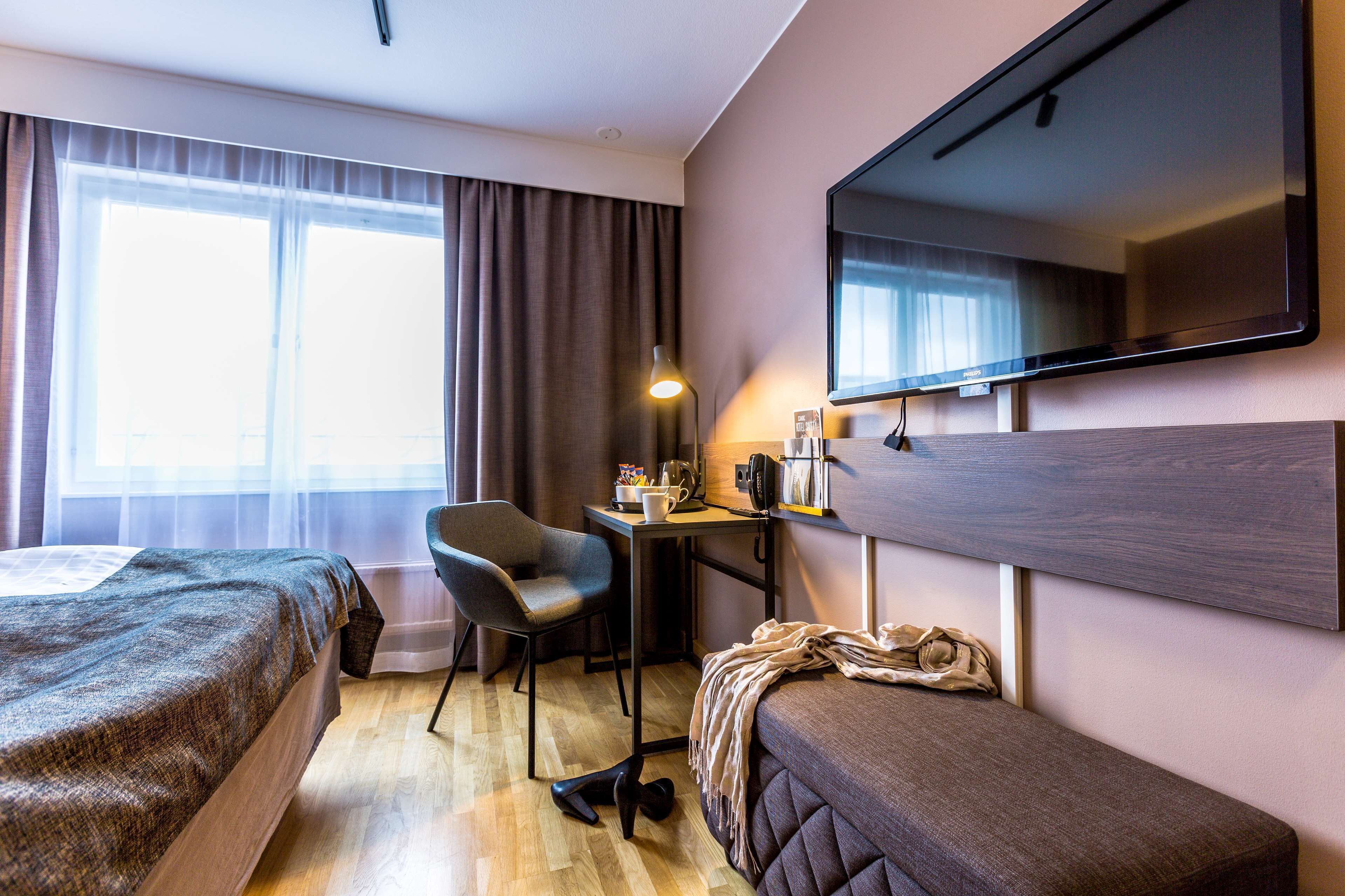 A hotel room with a bed, a desk, a chair, a TV, and a large window with curtains.
