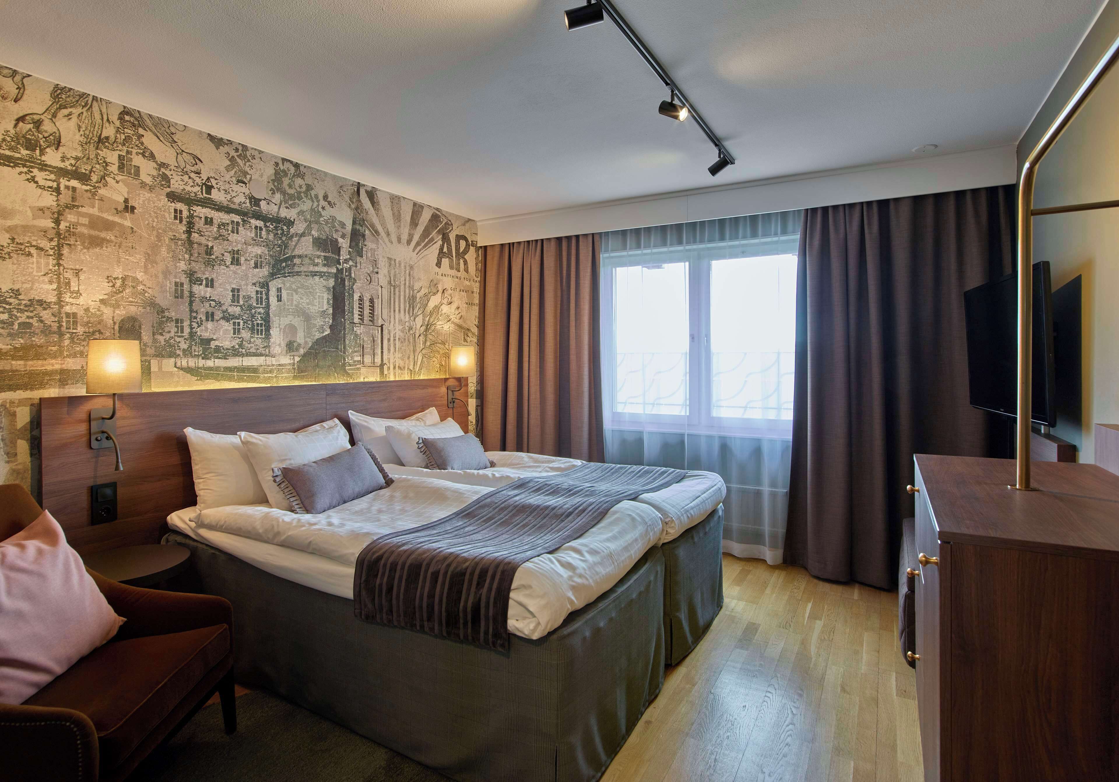 Une chambre d’hôtel avec un grand lit, une tête de lit ornée d’œuvres d’art, une télévision et une fenêtre avec des rideaux.