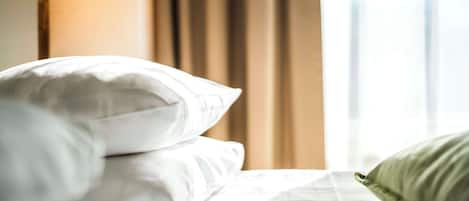 A neatly made bed with white bedding and a green pillow, a lamp, and a window with curtains in the background.