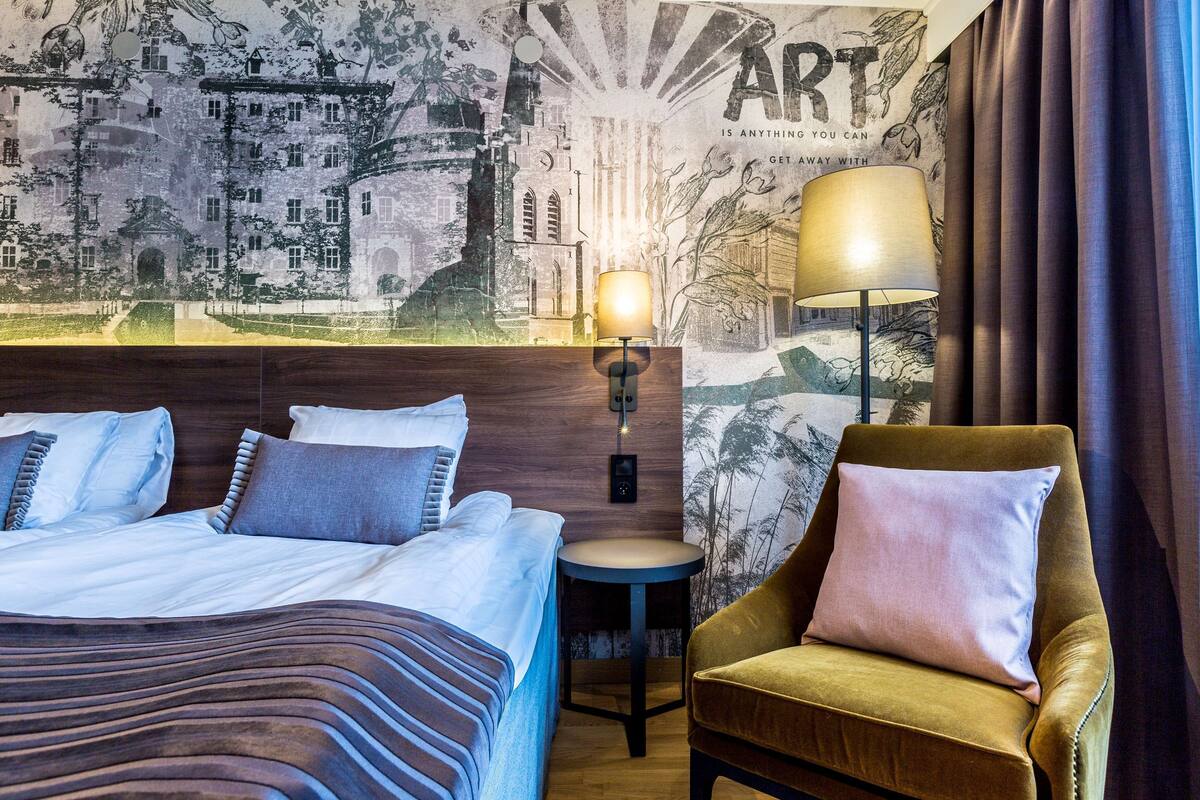 A hotel room with a bed, a chair, a wall-mounted lamp, and a mural.