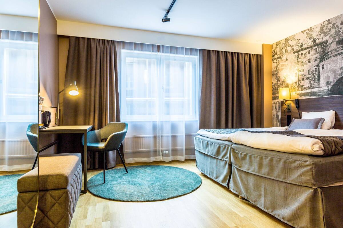 A hotel room with a bed, a desk, a chair, a small ottoman, a round rug, and a large window with curtains.