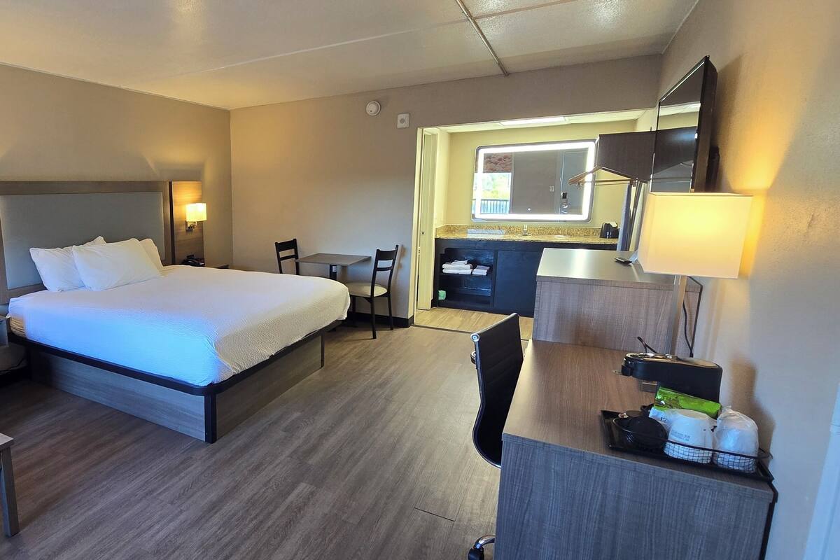 A hotel room with a bed, a sofa, a desk, a chair, a small table, a mirror, and a kitchenette area.