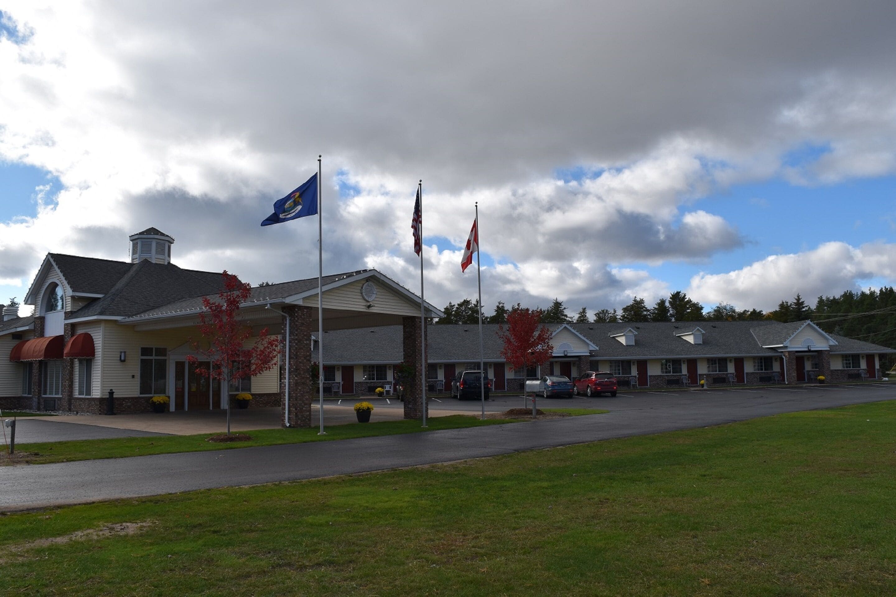 A motel with a covered parking area, multiple flags, and a row of buildings in the background.