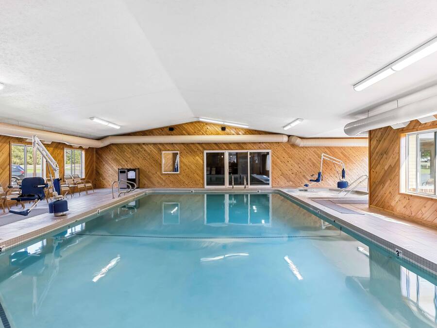 An indoor swimming pool with wooden walls, a clear pool, and a sunlit area with chairs and a ladder.