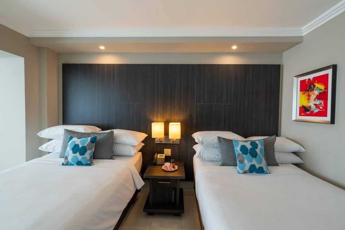 Quarto de hotel com duas camas, uma mesinha de cabeceira, duas luminárias fixadas na parede e uma obra de arte emoldurada.