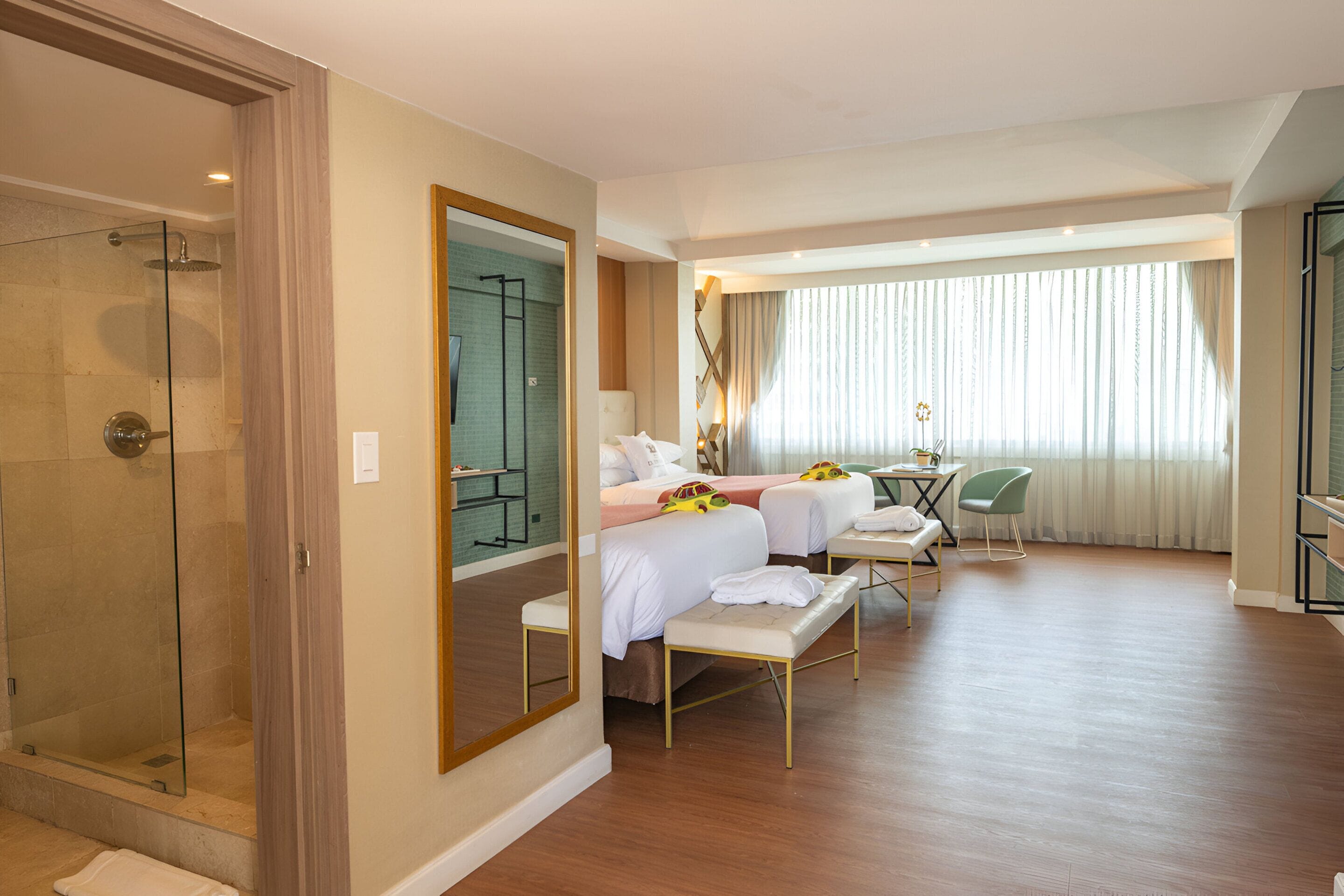 A hotel room with a glass-enclosed shower, two beds with white linens, a small table, a chair, and a large window with curtains.