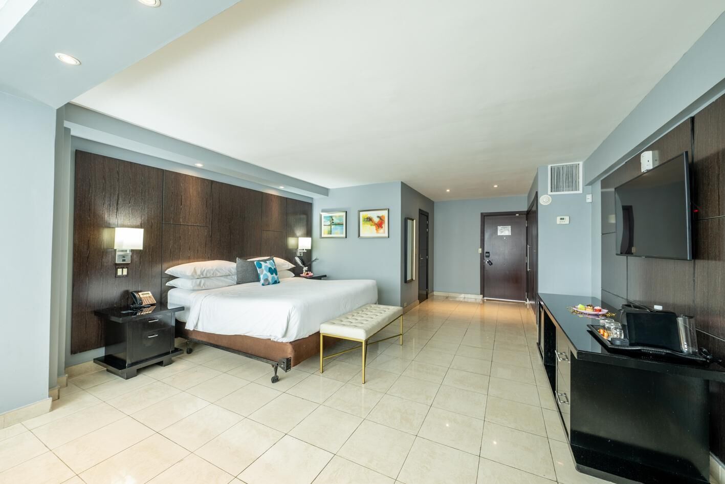 A hotel room with a bed, bedside table, lamp, artwork, and a kitchenette.
