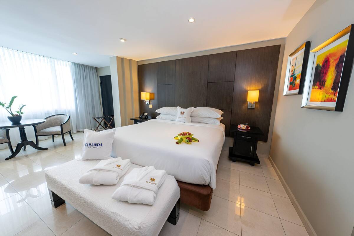 Una habitación de hotel con cama, mesitas de noche, escritorio, silla y ventana con cortinas.