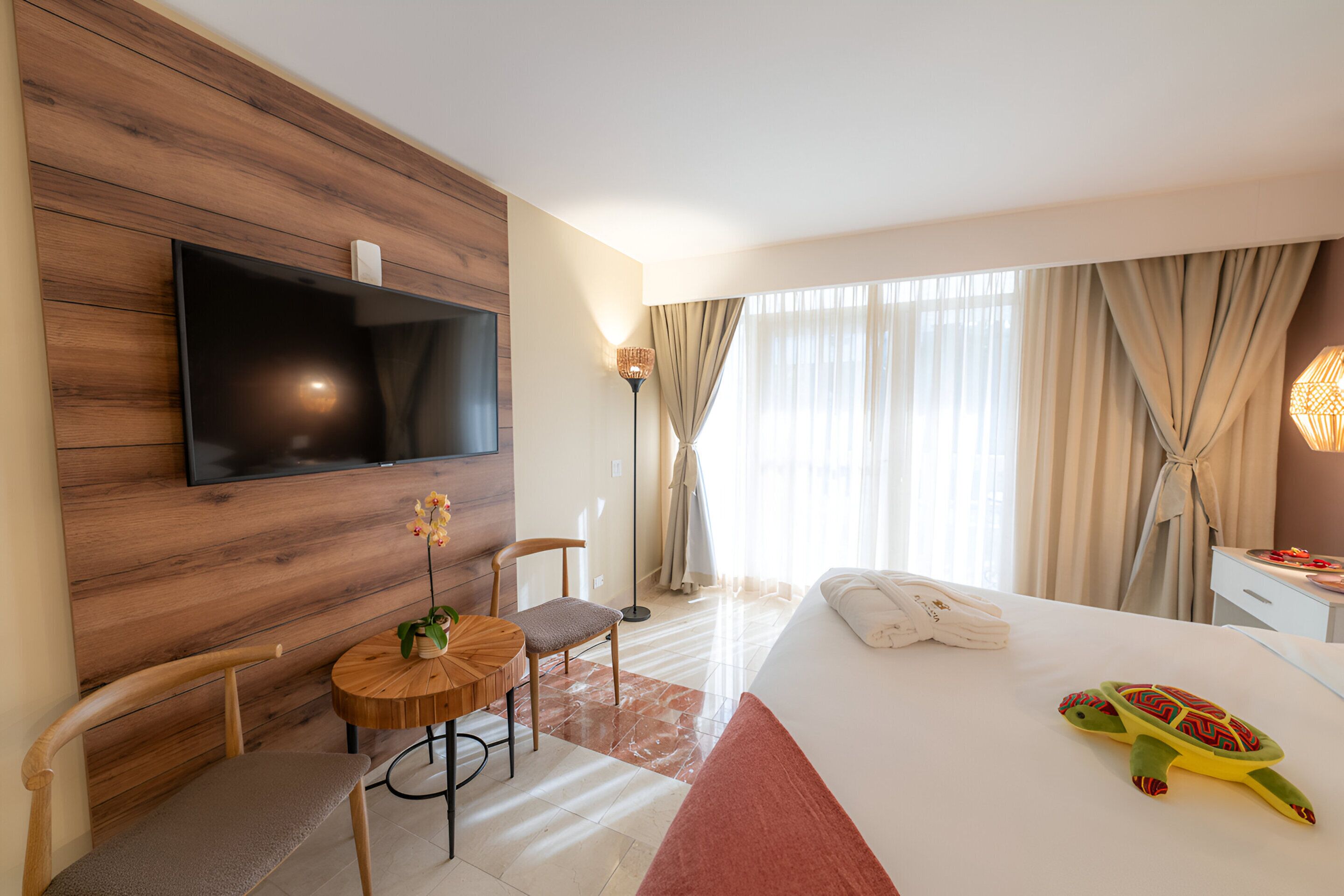 A hotel room with a wooden wall, a flat-screen TV, a round wooden table with a chair, a bed with a red accent, and a turtle plush toy on the bed.
