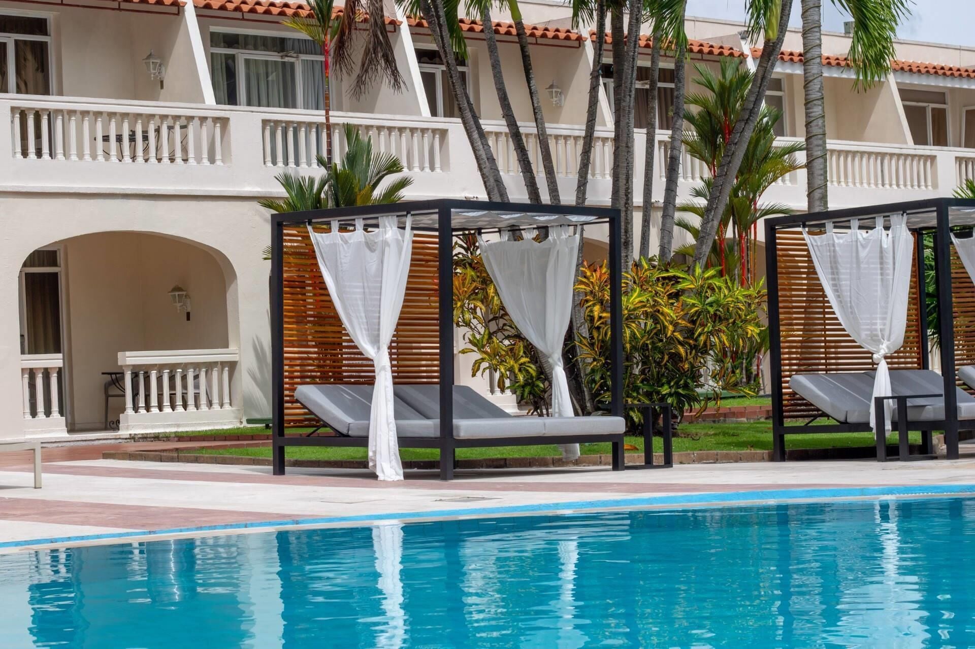 Un área junto a la piscina con sillones, una zona de estar techada y un edificio con tejado de azulejos y columnas.