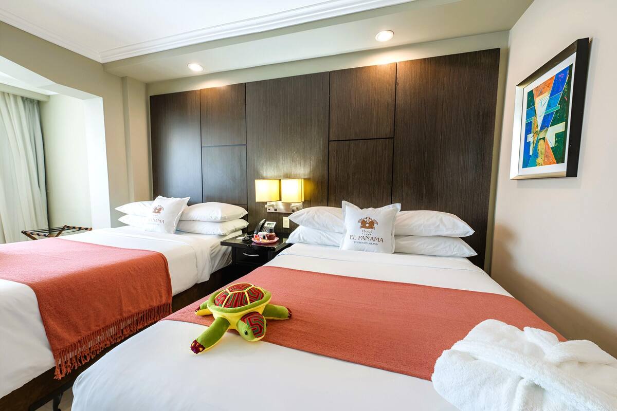 Habitación de hotel con dos camas, un peluche, una lámpara y un cuadro en la pared.