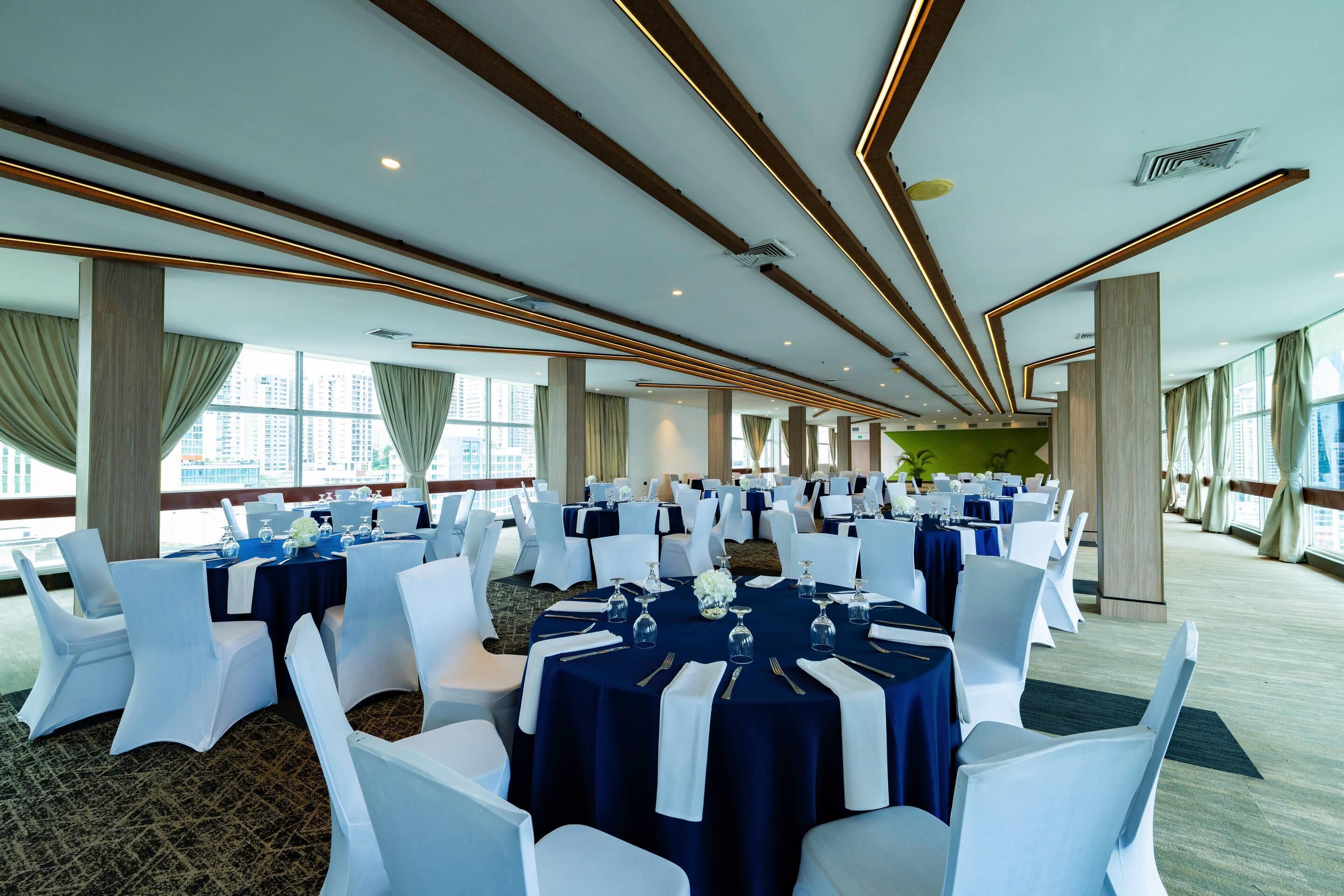 Une salle de banquet avec des tables rondes disposées pour les événements, dotée d’un plafond moderne avec des poutres en bois et un éclairage encastré.