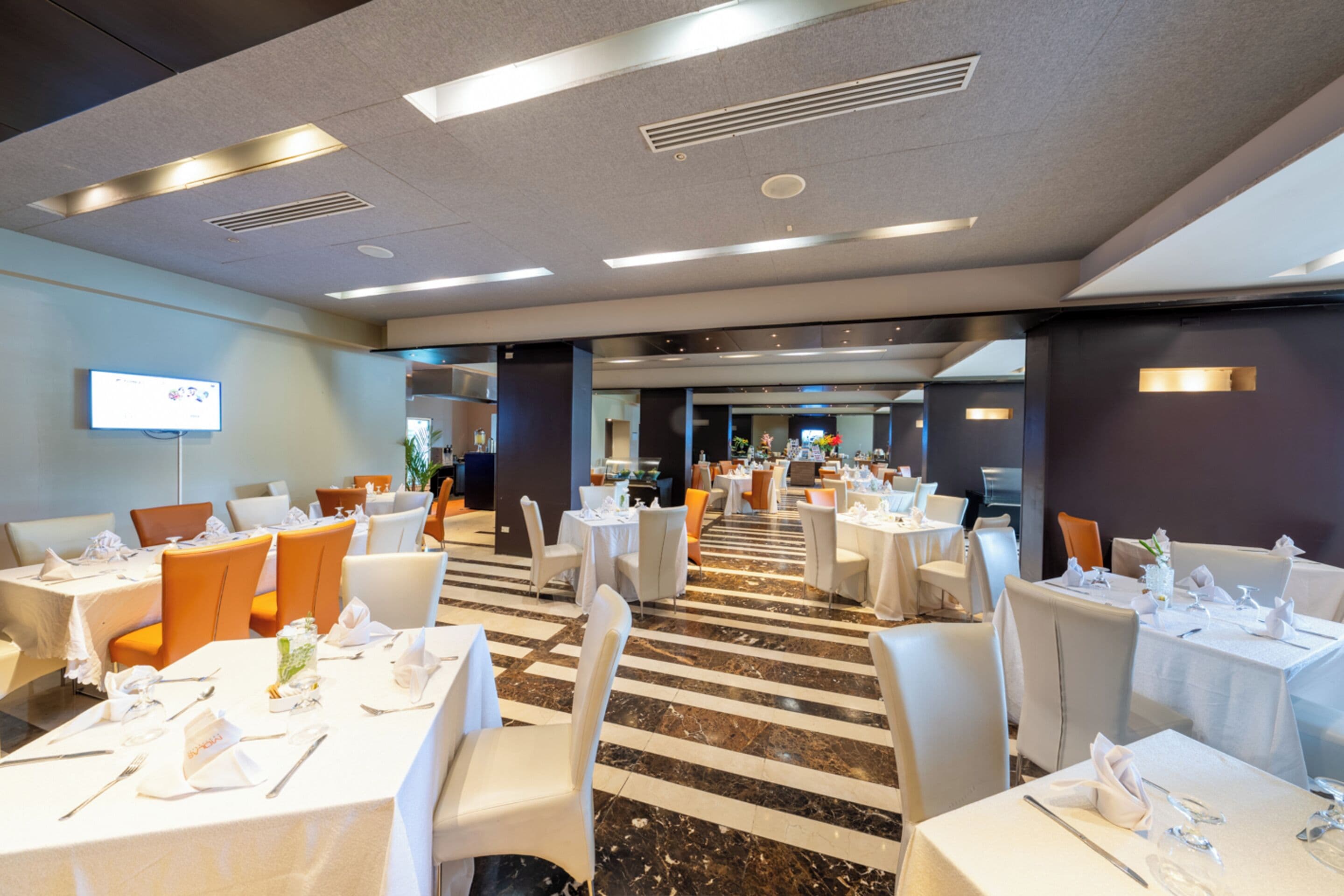 A spacious hotel dining area with multiple tables and chairs, a central walkway, and a television screen on the wall.