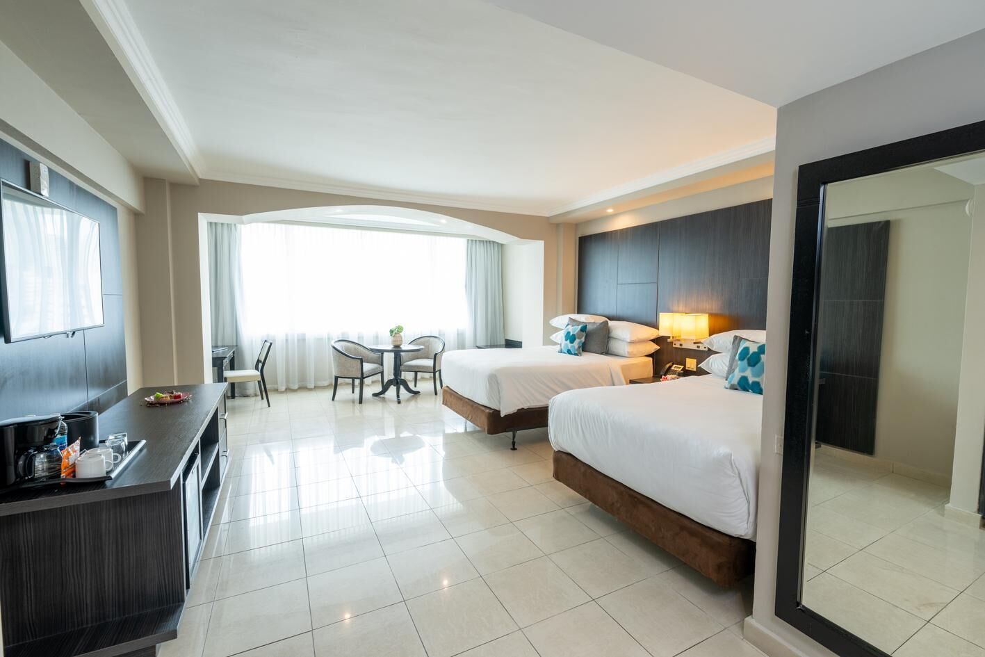 Quarto de hotel com duas camas, uma escrivaninha com televisão, zona de refeições com cadeiras e uma janela grande.