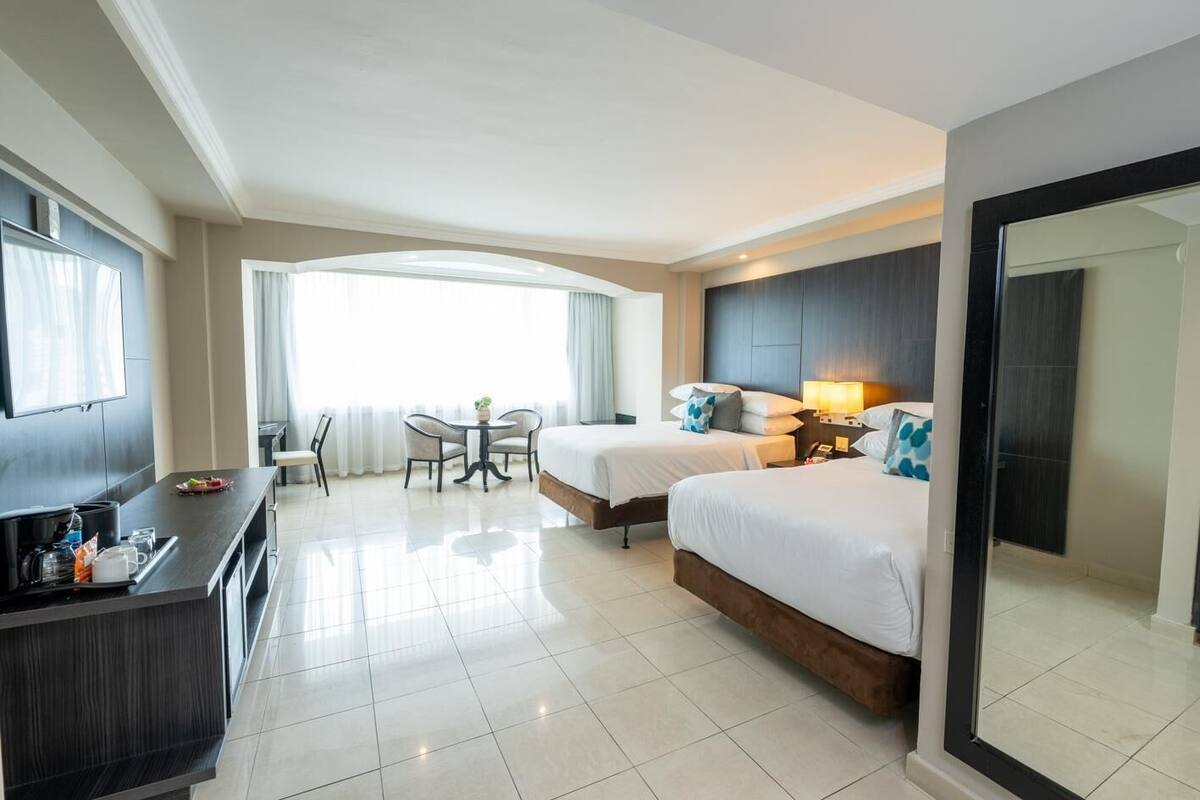 Habitación de hotel con dos camas, un escritorio con televisor, una zona de comedor con sillas y un ventanal grande.