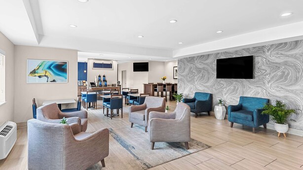 Un moderno vestíbulo de hotel con sillones tapizados en gris y azul, una pared grande con diseño en gris y un televisor de pantalla plana sobre un mostrador de recepción.