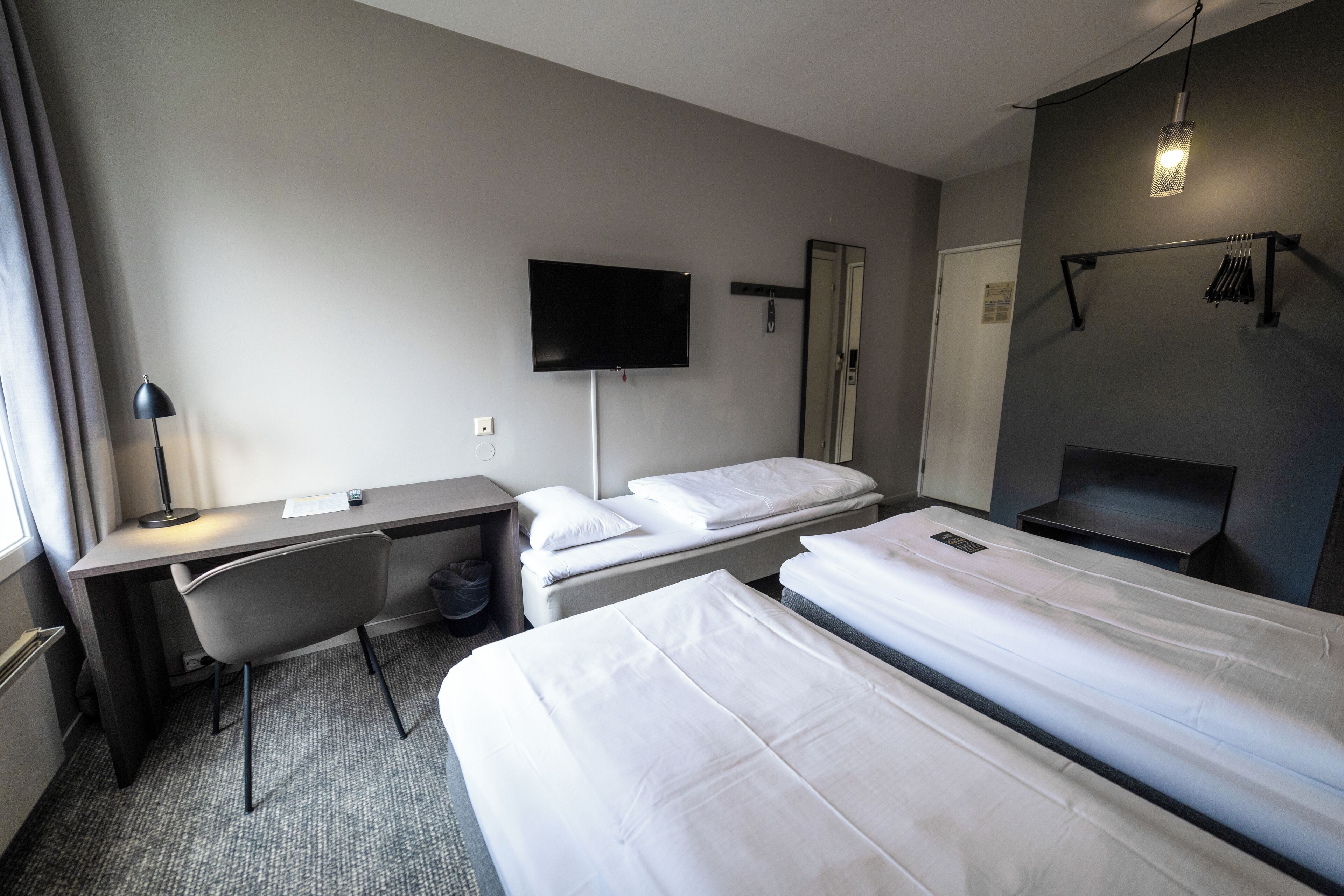 Une chambre d’hôtel avec deux lits simples, un bureau avec une chaise, un téléviseur à écran plat fixé au mur et un petit coin salon avec un banc et une table.