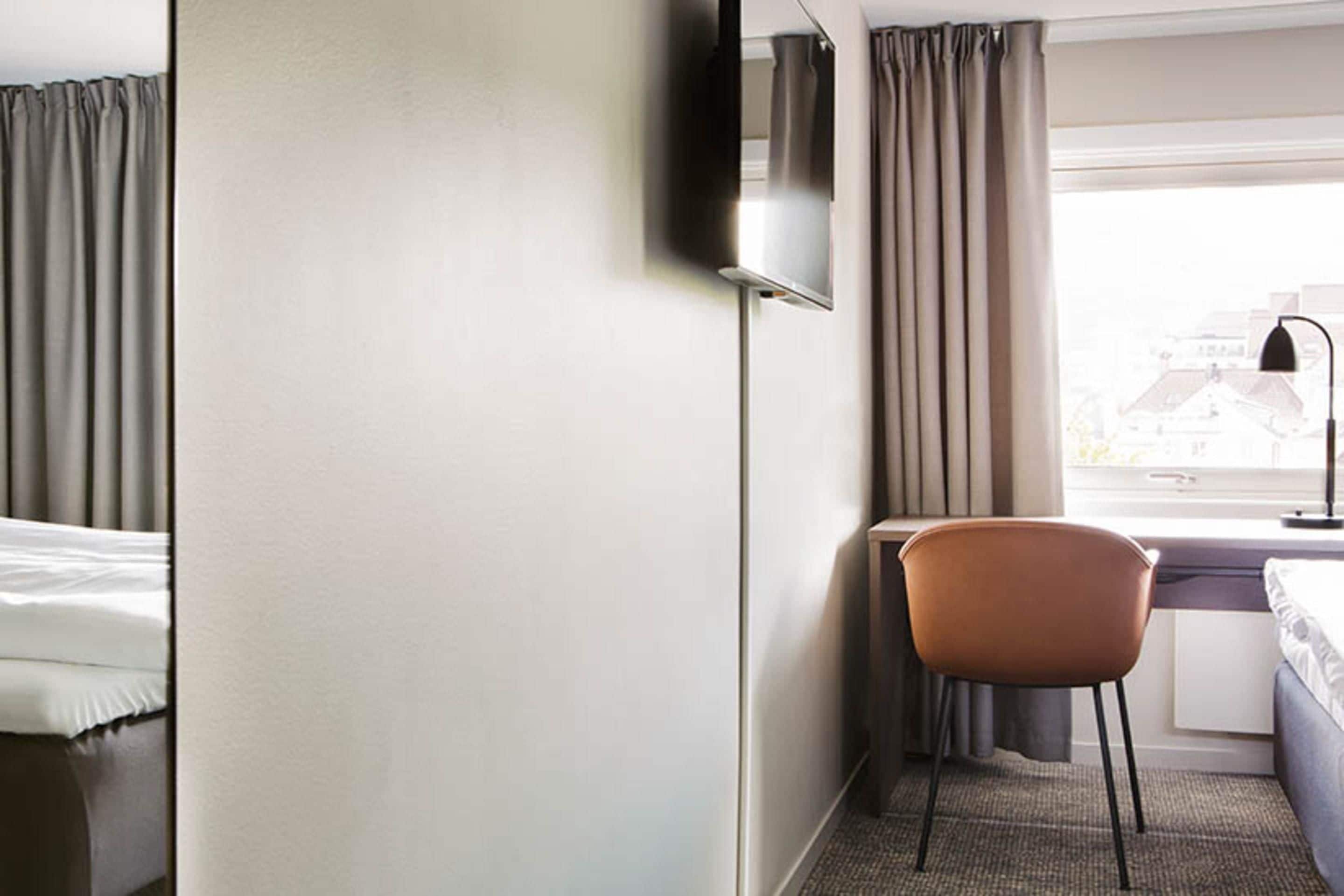 Een hotelkamer met een bed, een bureau, een stoel, een aan de muur bevestigde televisie en een raam met gordijnen.