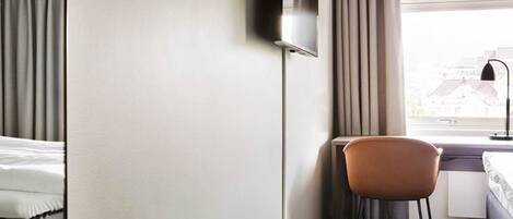 Habitación de hotel con cama, escritorio, silla, televisor montado en la pared y ventana con cortinas.