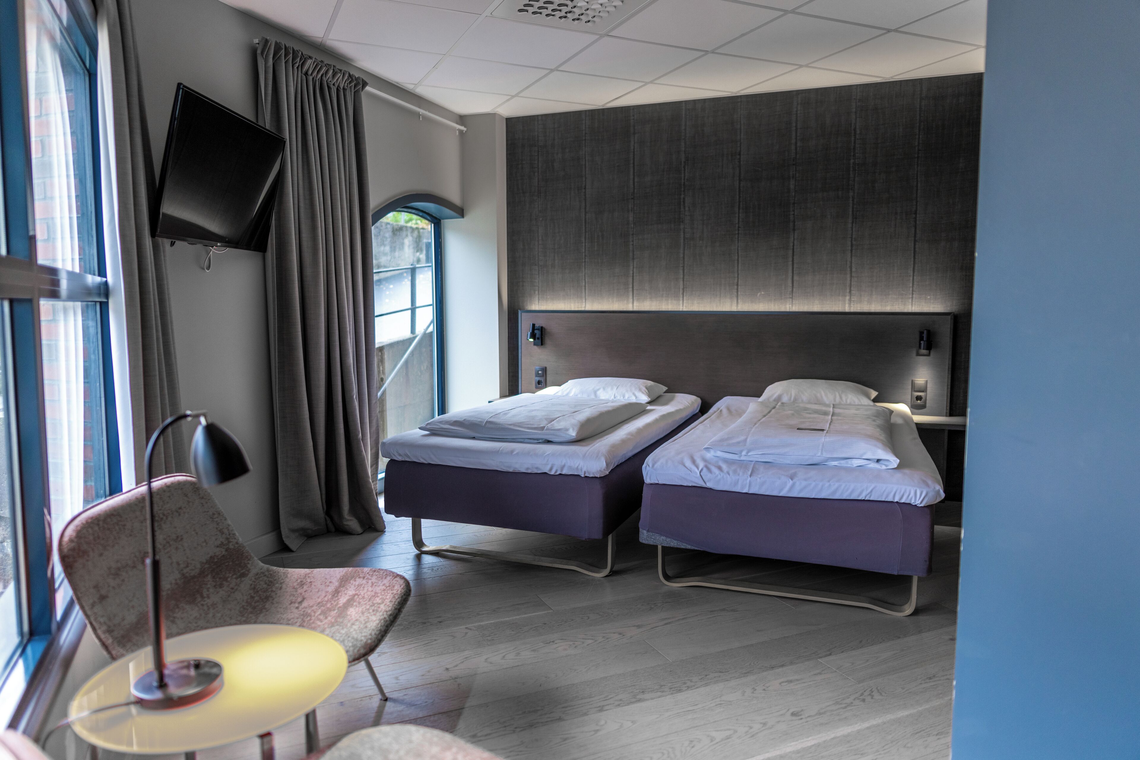 A modern hotel room with two beds, a desk, a chair, a TV, and a window with curtains.