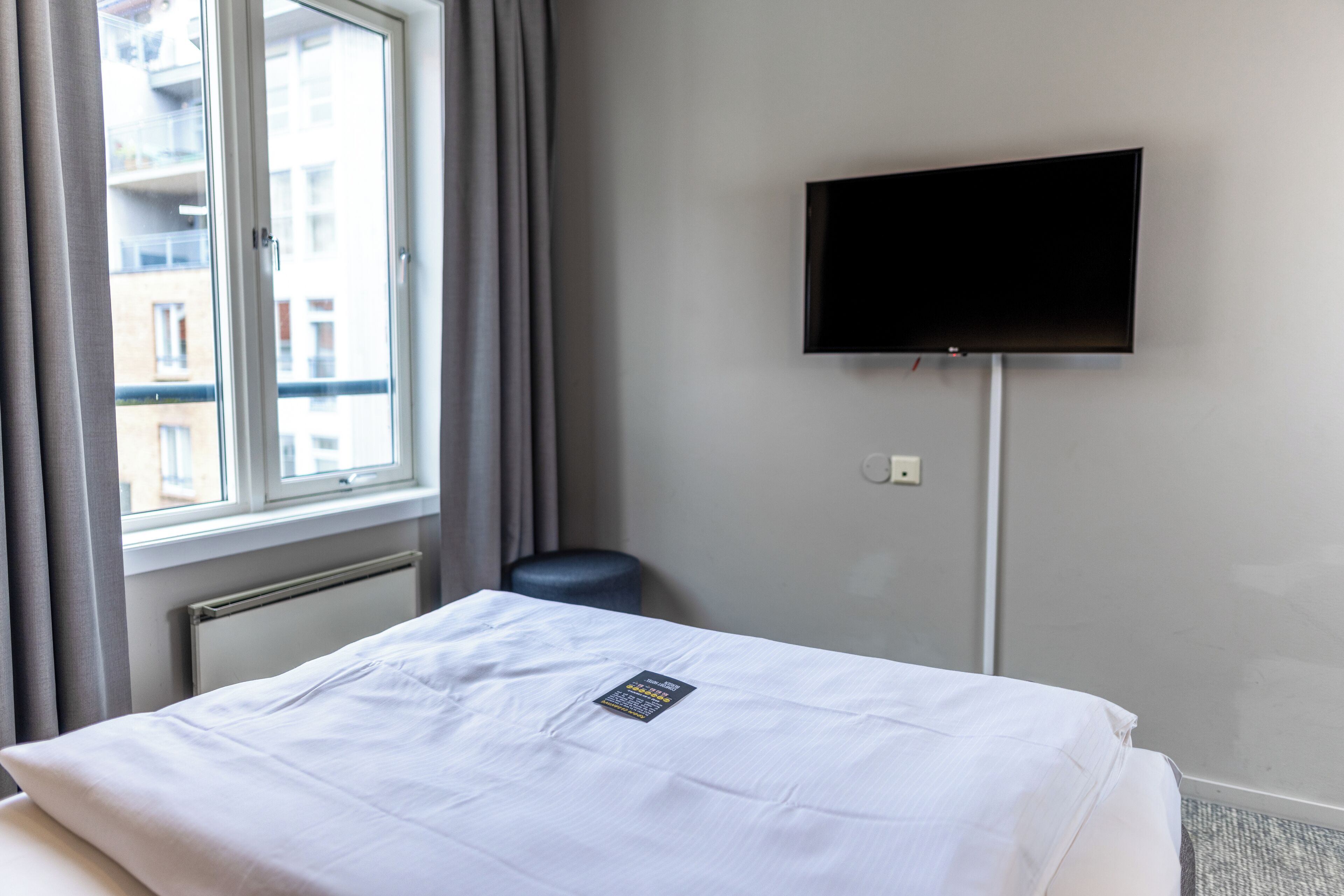 A hotel room with a bed, a TV mounted on the wall, a window with curtains, and a radiator.