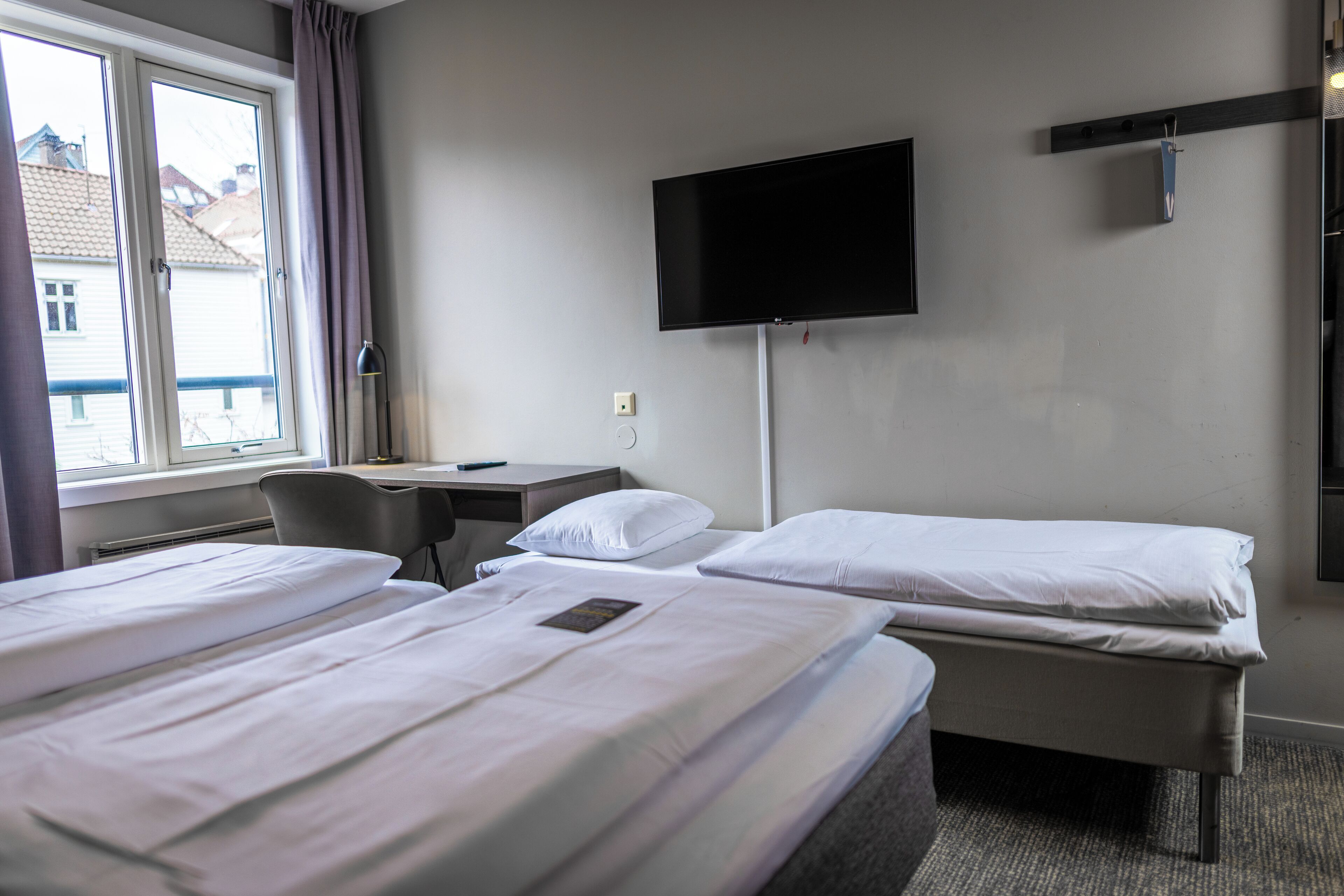 A hotel room with two beds, a desk, a chair, a TV mounted on the wall, and a window with curtains.