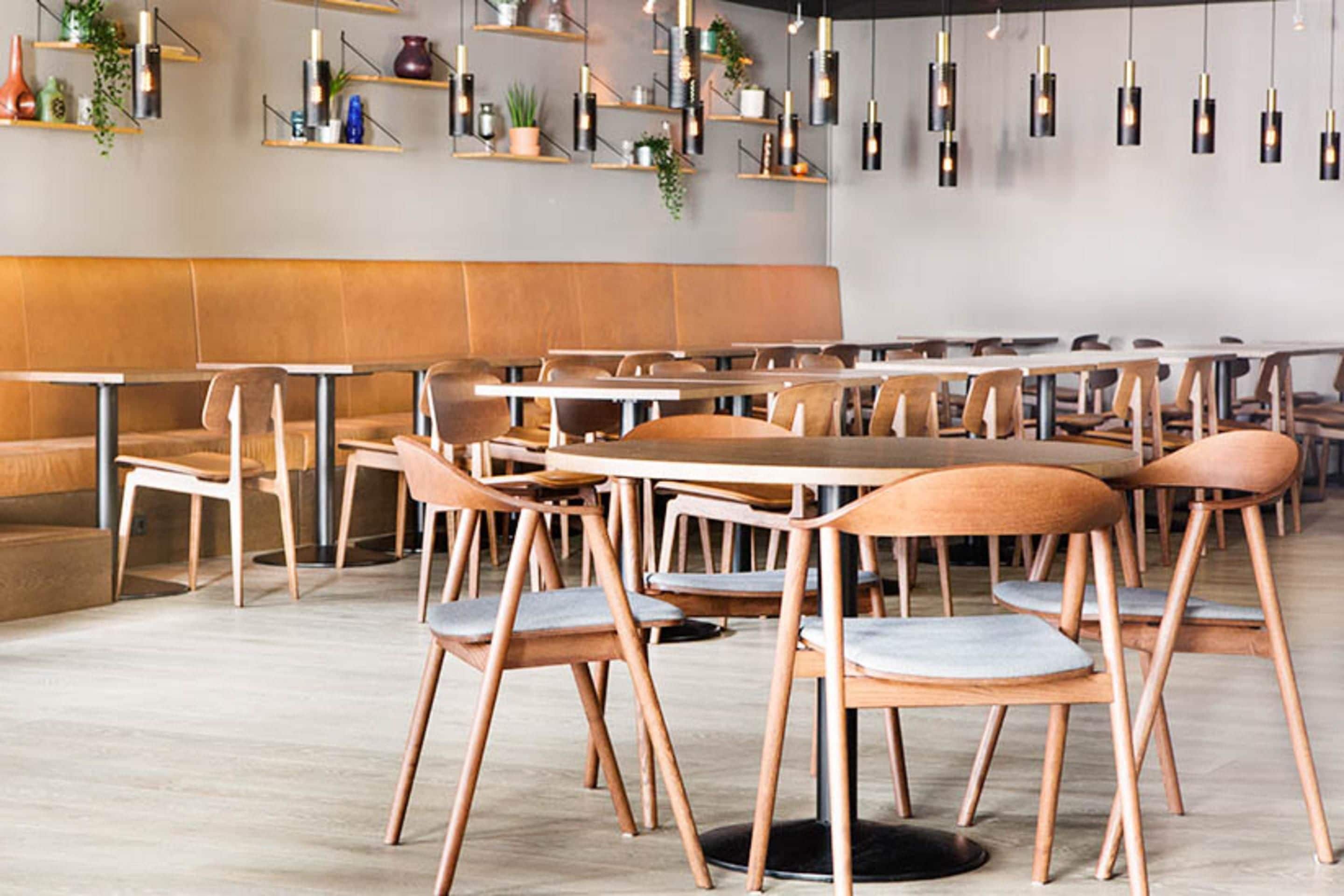 Un interior de restaurante moderno con mesas y sillas de madera, un banco largo e iluminación colgante.