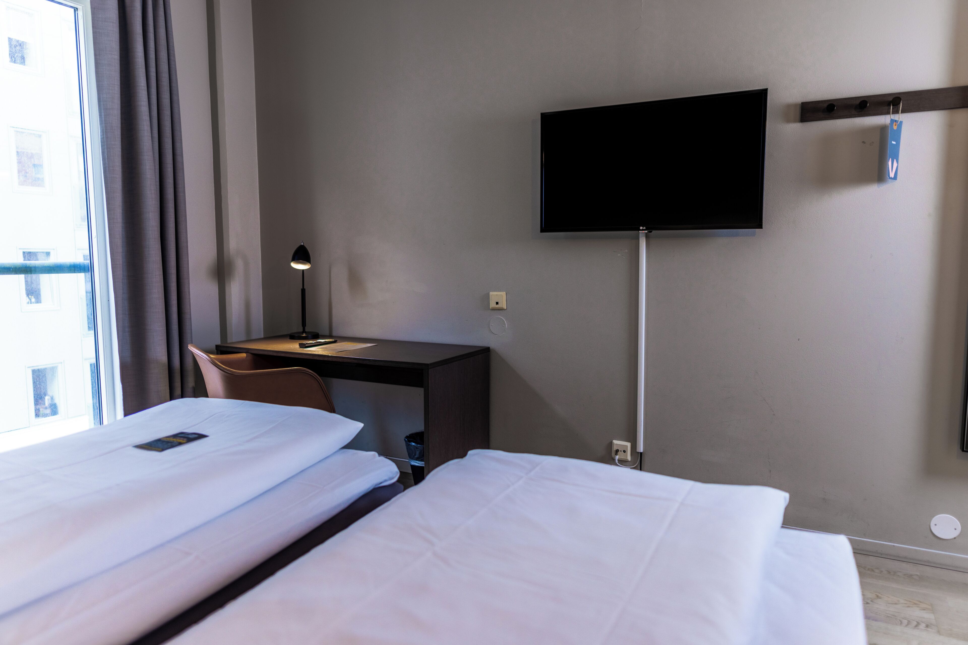 Ein Hotelzimmer mit zwei Betten, einem Schreibtisch, einem Stuhl, einem flachbildfernseher an der Wand und einem Fenster mit Vorhängen.