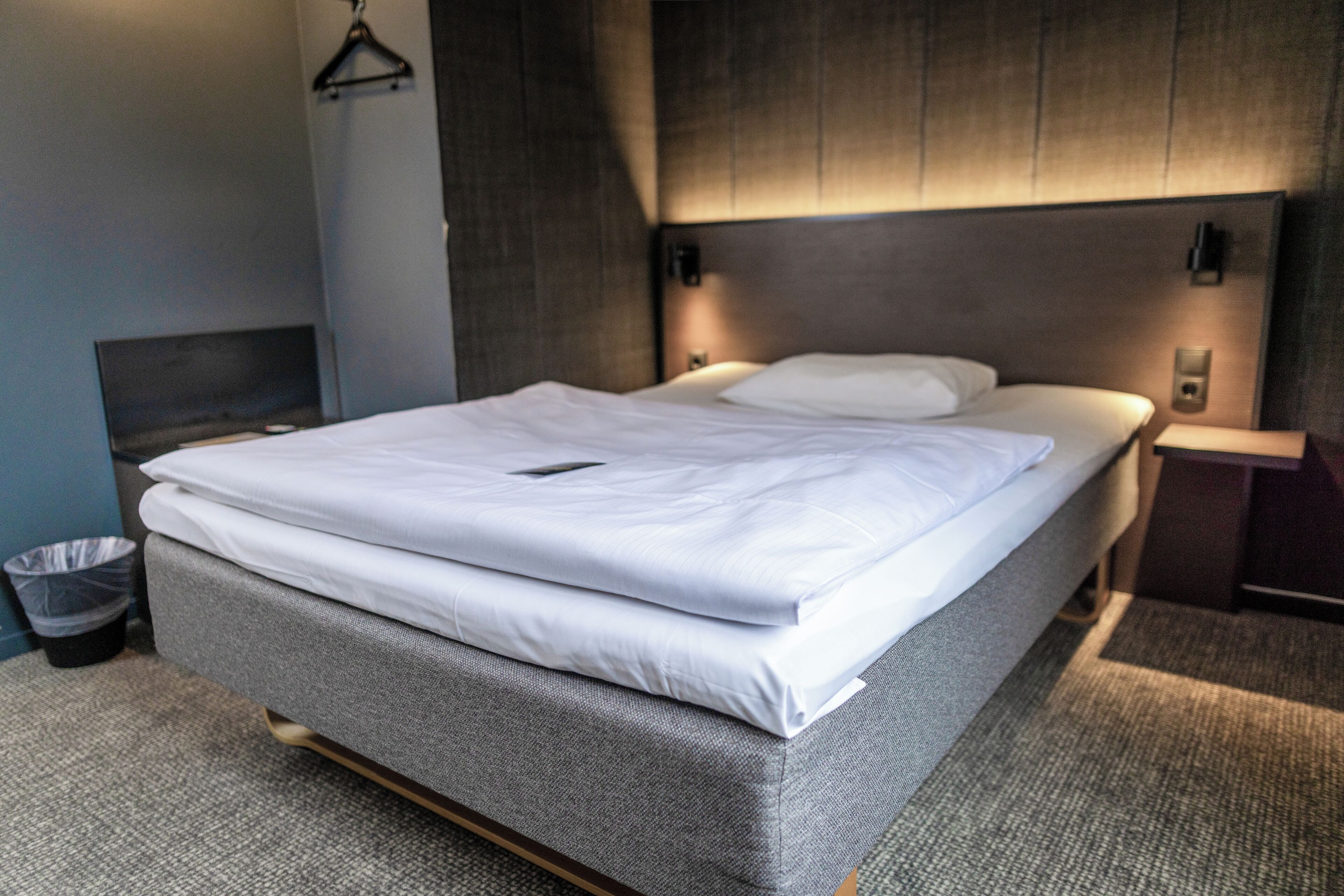 A neatly made bed with white linens, a grey upholstered headboard, and a small bedside table with a lamp.
