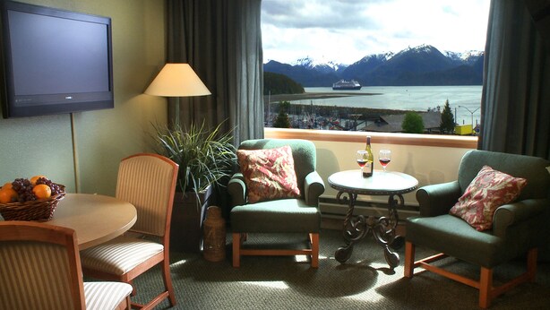Une chambre d’hôtel équipée d’une télévision, de deux fauteuils, d’une petite table avec des verres à vin, d’un panier de fruits et offrant une vue sur les montagnes et un navire.