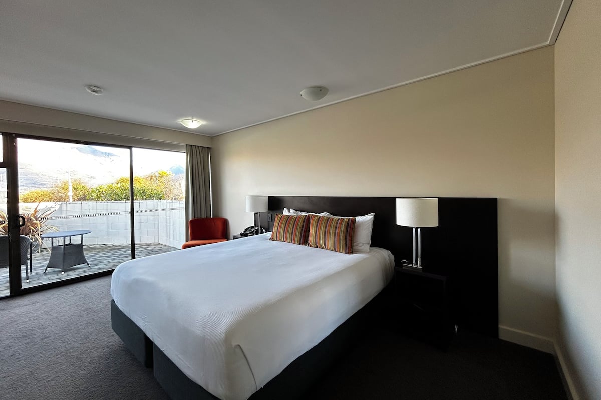 Une chambre d’hôtel avec un grand lit, une table de chevet, une chaise et une vue sur l’extérieur grâce à une porte-fenêtre coulissante.