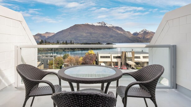 A balcony with wicker furniture and a glass table overlooking a mountain and lake.