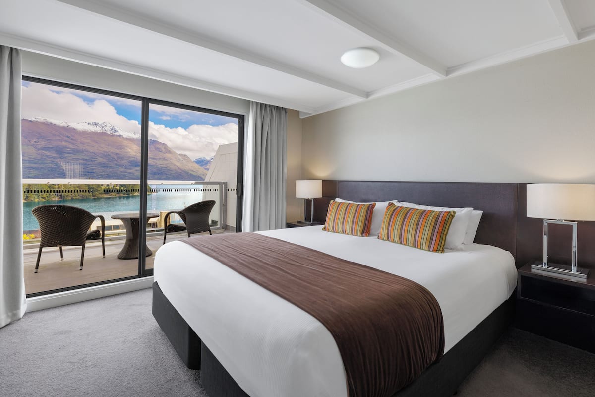 Une chambre d’hôtel avec un grand lit, une vue sur un lac et des montagnes, un balcon avec des chaises et une table de chevet avec une lampe.