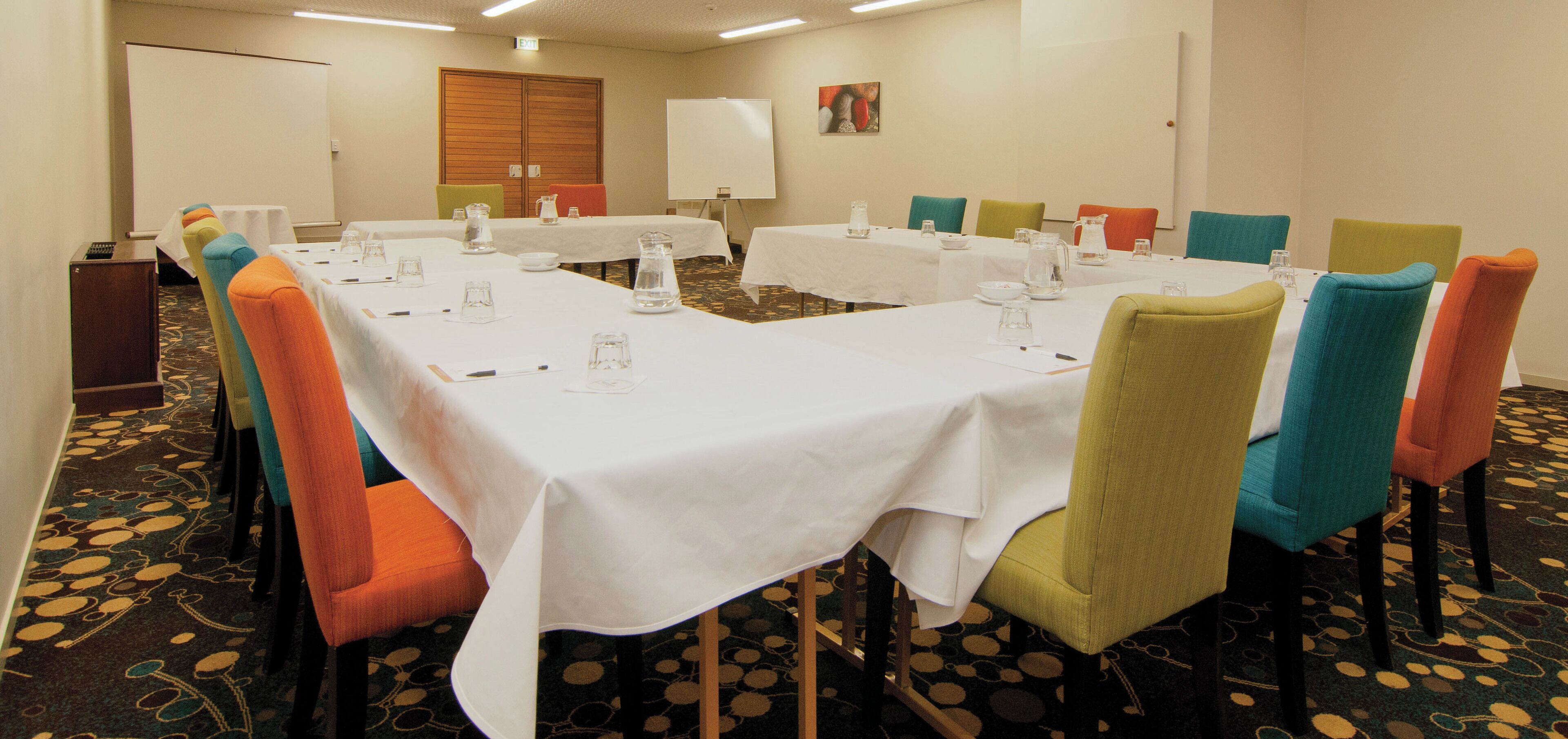 A conference room with multiple tables and chairs, equipped with water pitchers and glasses, and a projector screen.