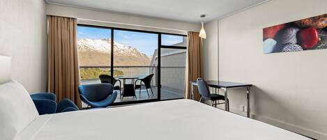 Habitación de hotel con una cama grande, un escritorio, dos sillas y vistas a montañas a través de un ventanal.