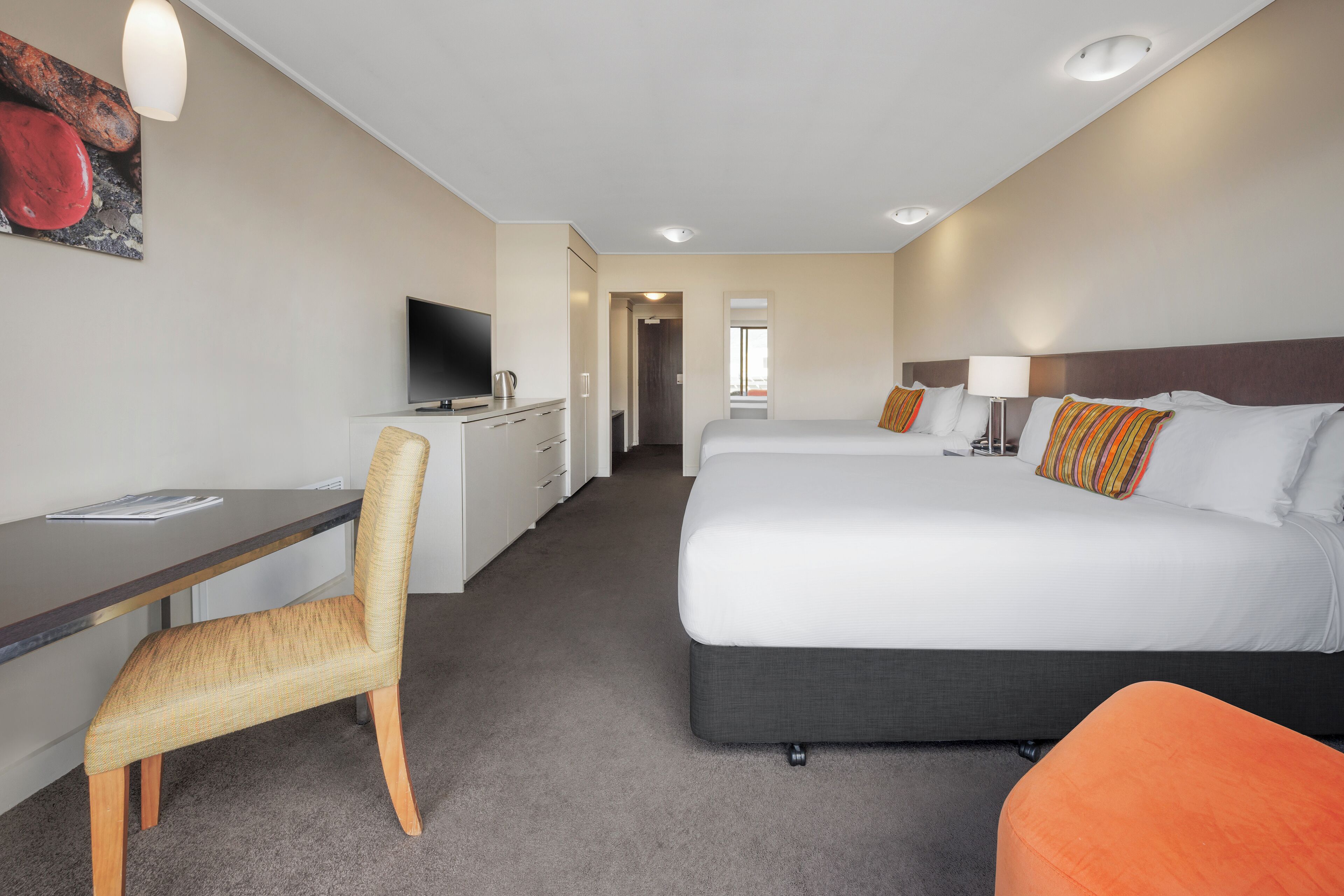 A hotel room with a desk, chair, two beds, a television, and a painting on the wall.