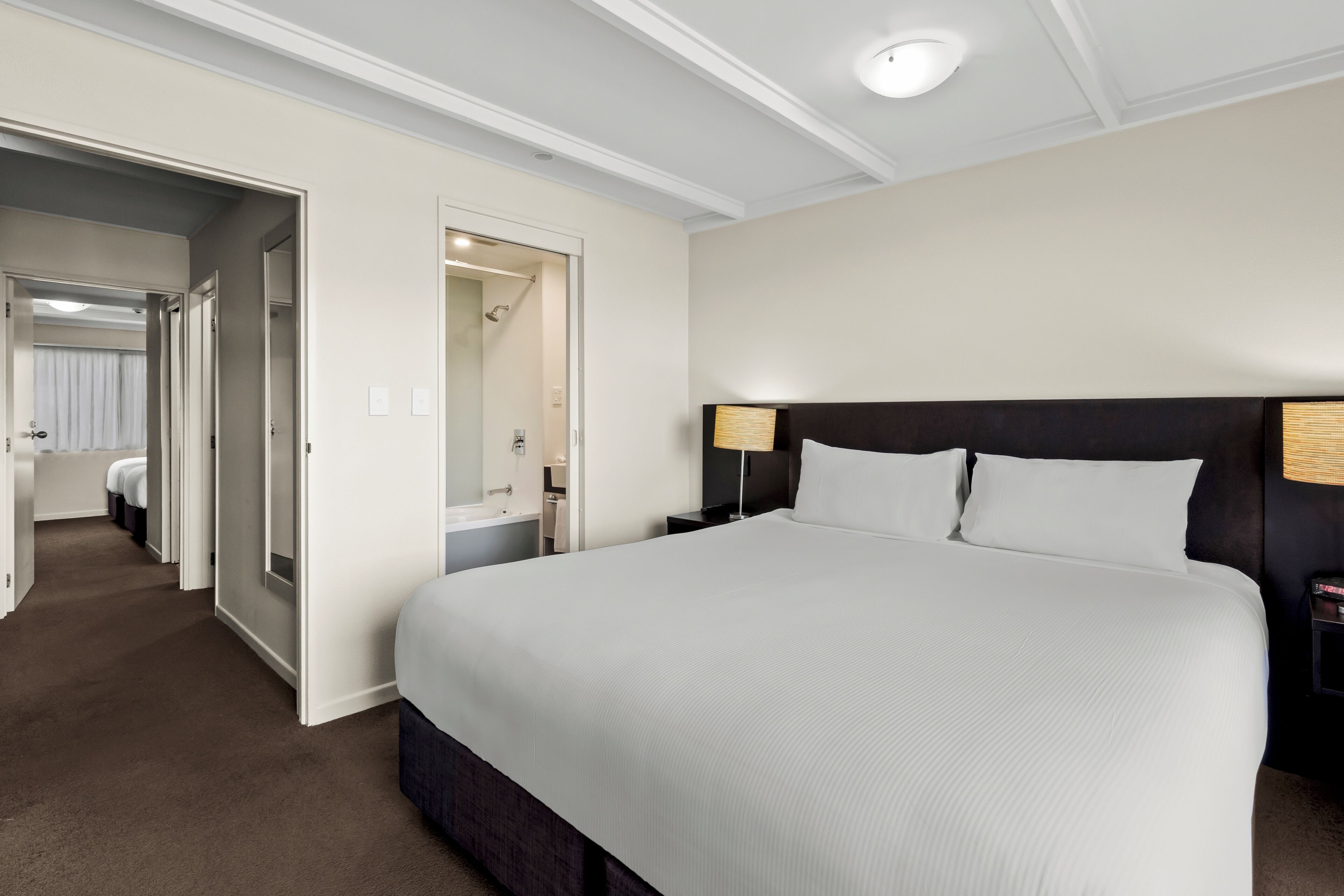 Une chambre d’hôtel avec un grand lit, une table de chevet, une lampe et une salle de bain visible à travers une porte ouverte.