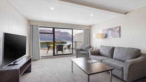 A modern hotel room with a flat-screen TV, a grey sofa, a coffee table, and a balcony with a mountain view.