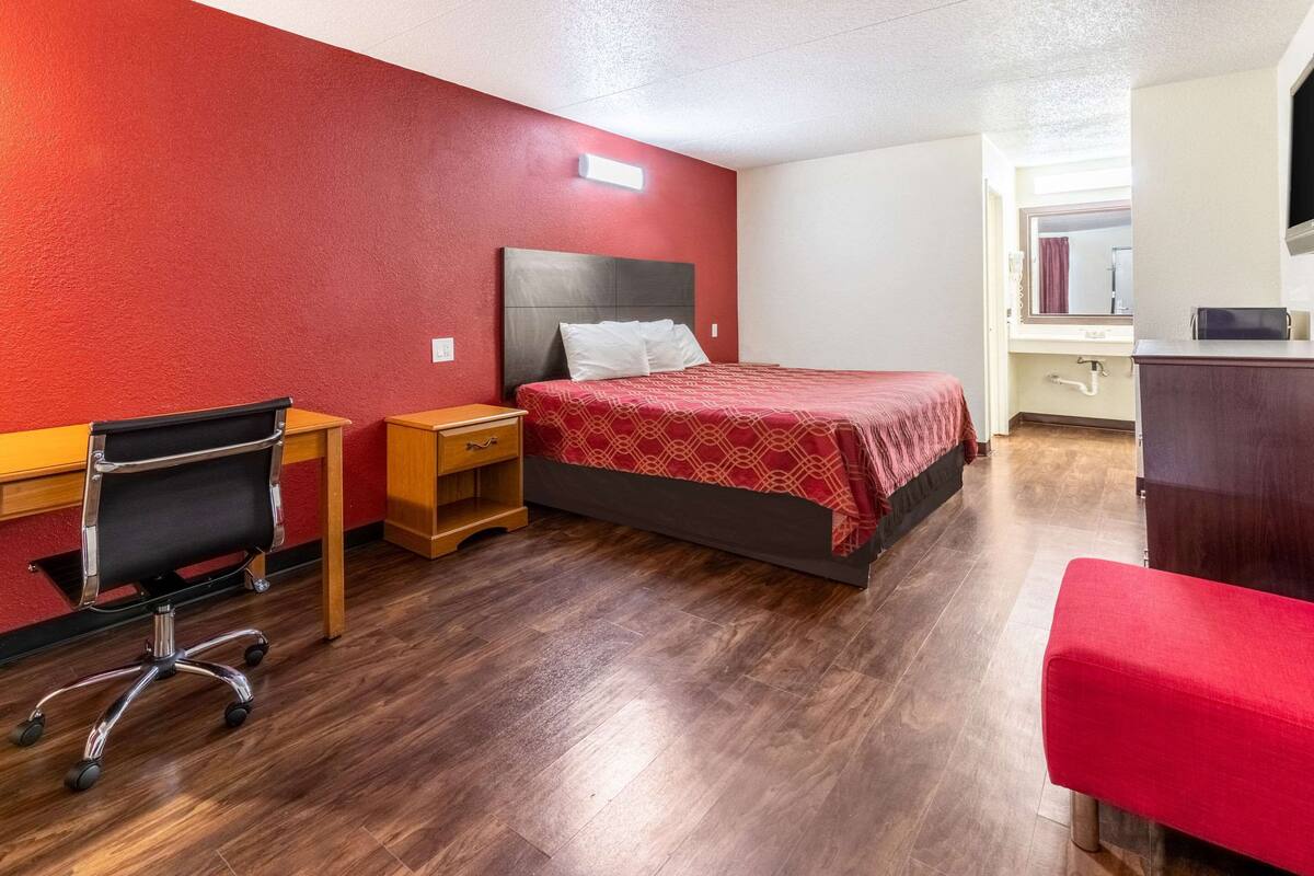 A hotel room with a bed, a desk with a chair, a small table, and a bench.