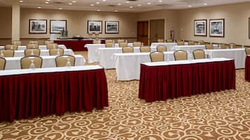 A conference room with rows of tables, each with a white tablecloth and red table runners. The room has a patterned carpet and beige walls.