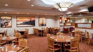 A hotel dining area with multiple wooden tables and chairs, a patterned carpet, and wall-mounted artwork depicting coastal scenes. - Clarion Hotel Portland Airport ME (Portland)