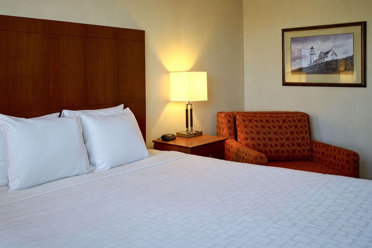 A hotel room with a large bed, a nightstand with a lamp, an orange chair, and a framed picture on the wall.