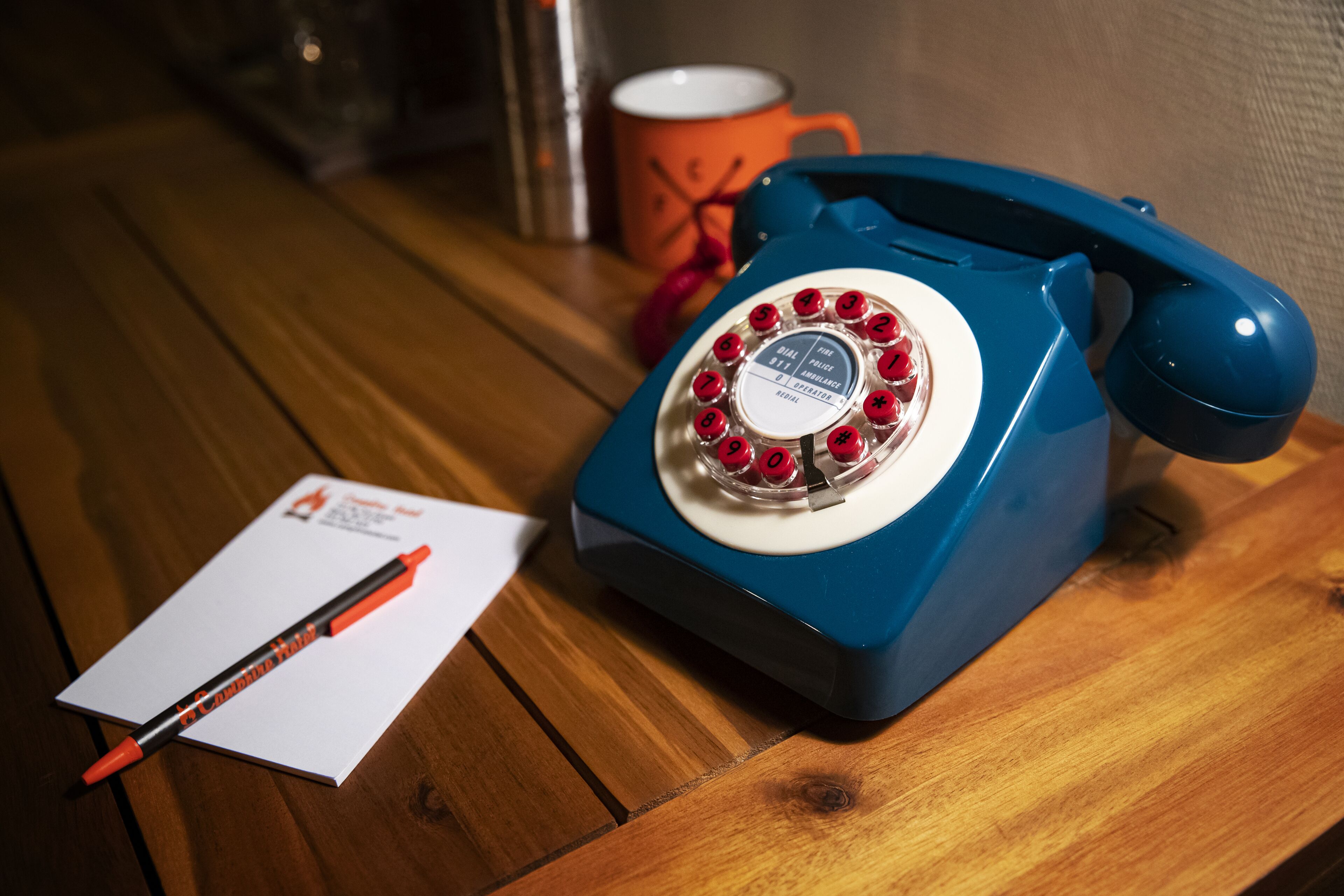 A blue rotary phone on a wooden table with a red pen and a piece of paper.