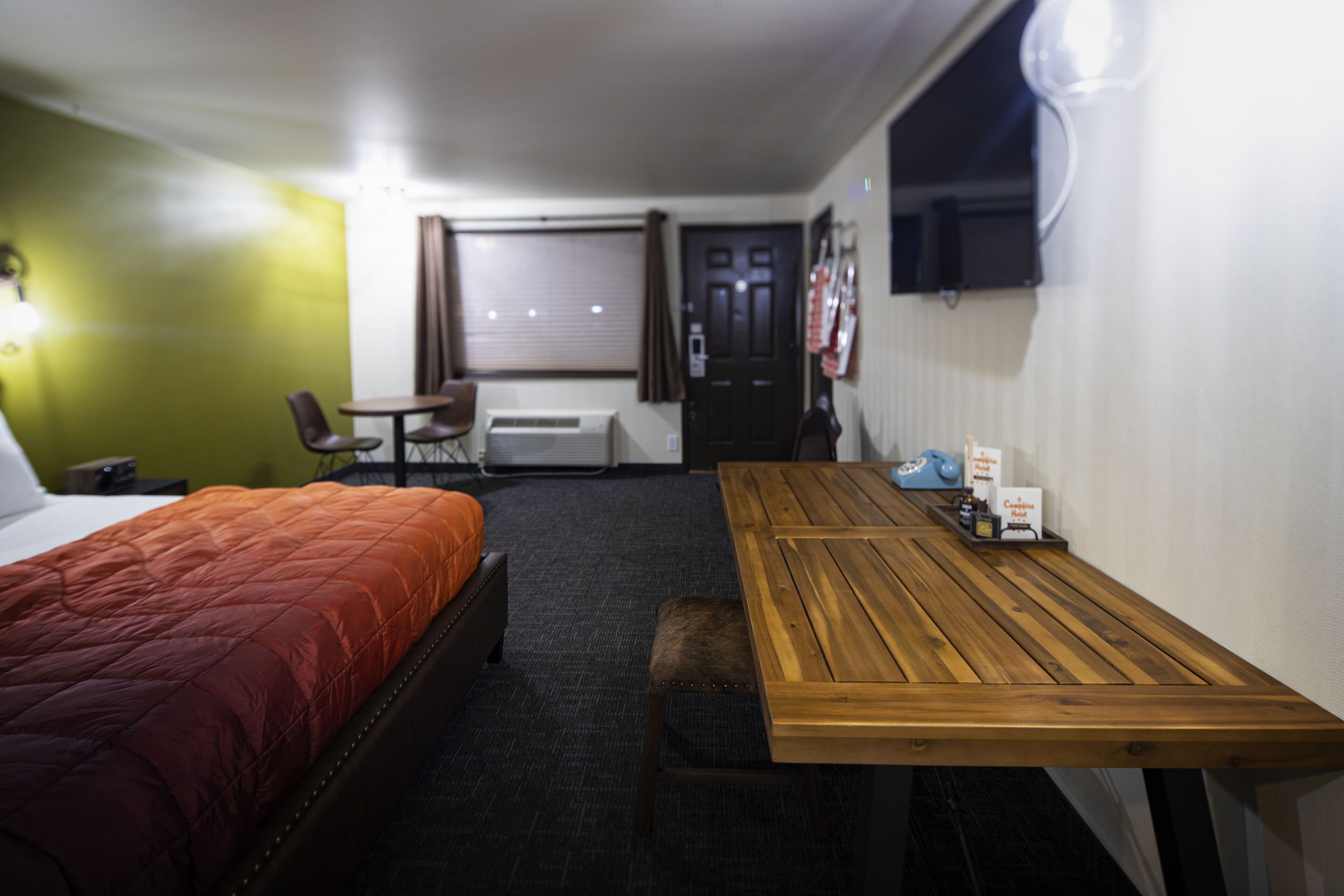 A hotel room with a bed, a wooden table, a chair, a small table, and a wall-mounted TV.