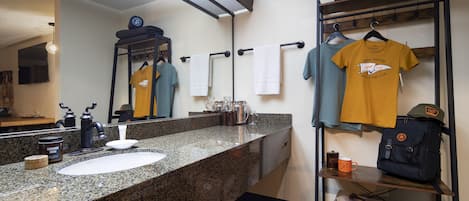 Un baño con lavamanos de encimera de granito, espejo, toallas y un estante con bolsas y una camiseta.