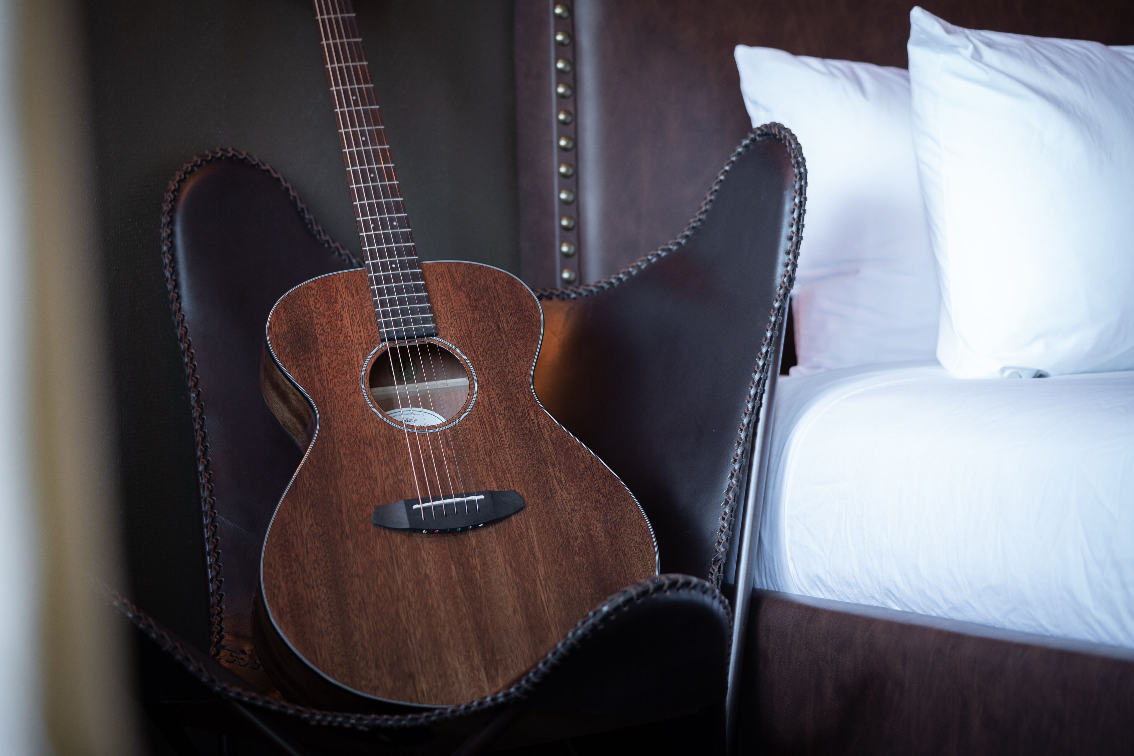 A guitar leaning against a dark leather chair in a hotel room with a bed and white pillows.