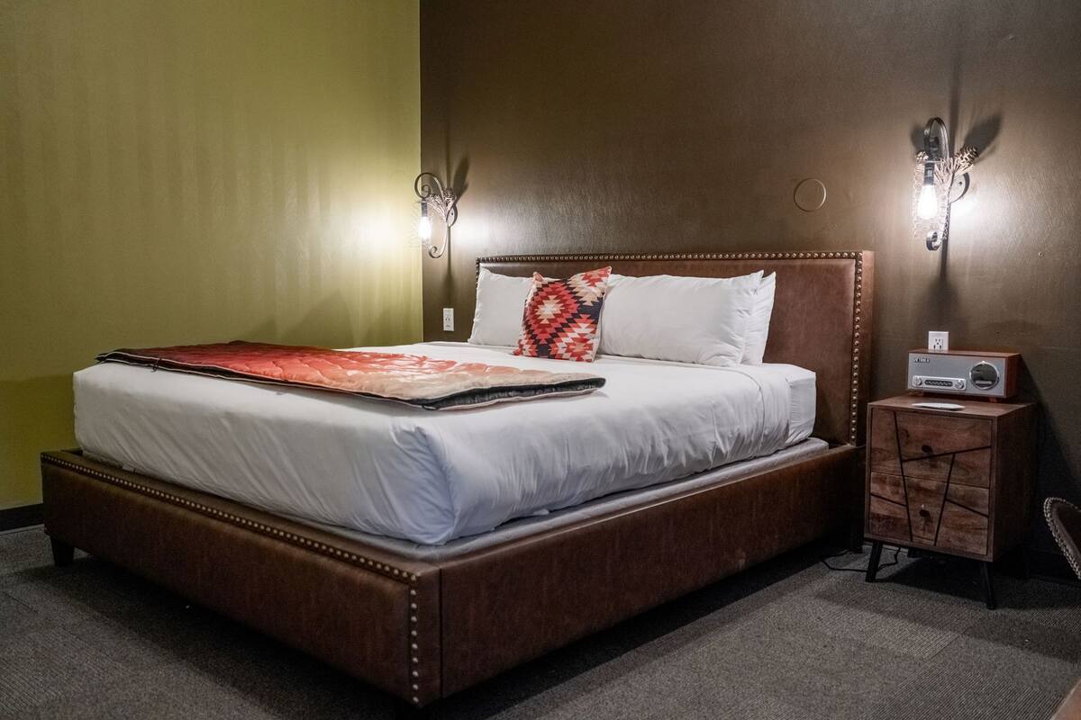 Una cama con una colcha roja, almohadas blancas y un cabecero de madera. Hay una mesita de noche con una radio y una lámpara.