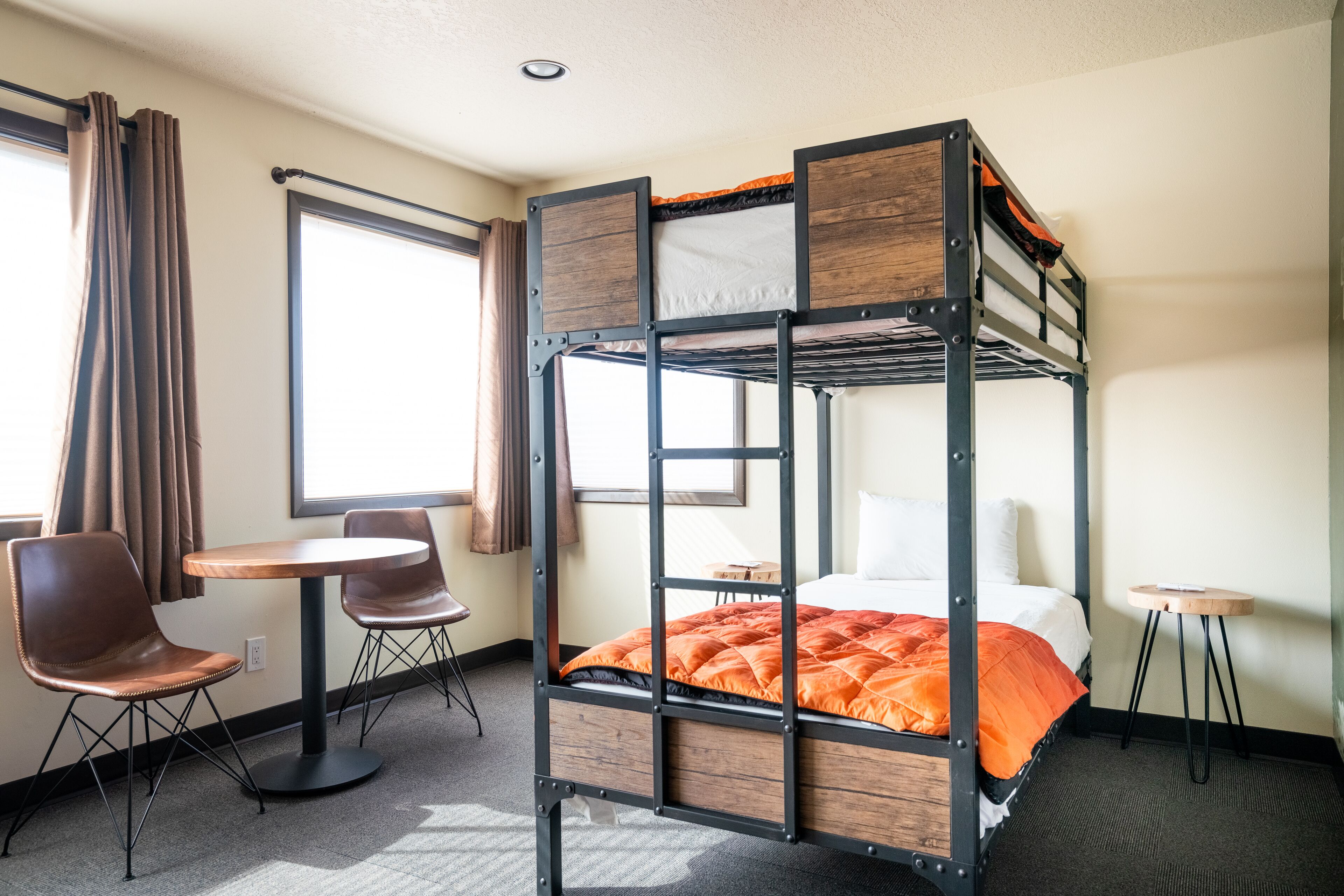 Ein Stockbett mit Holzbettrahmen und orangefarbener Bettwäsche, ein runder Tisch mit zwei Stühlen und ein kleiner Hocker.