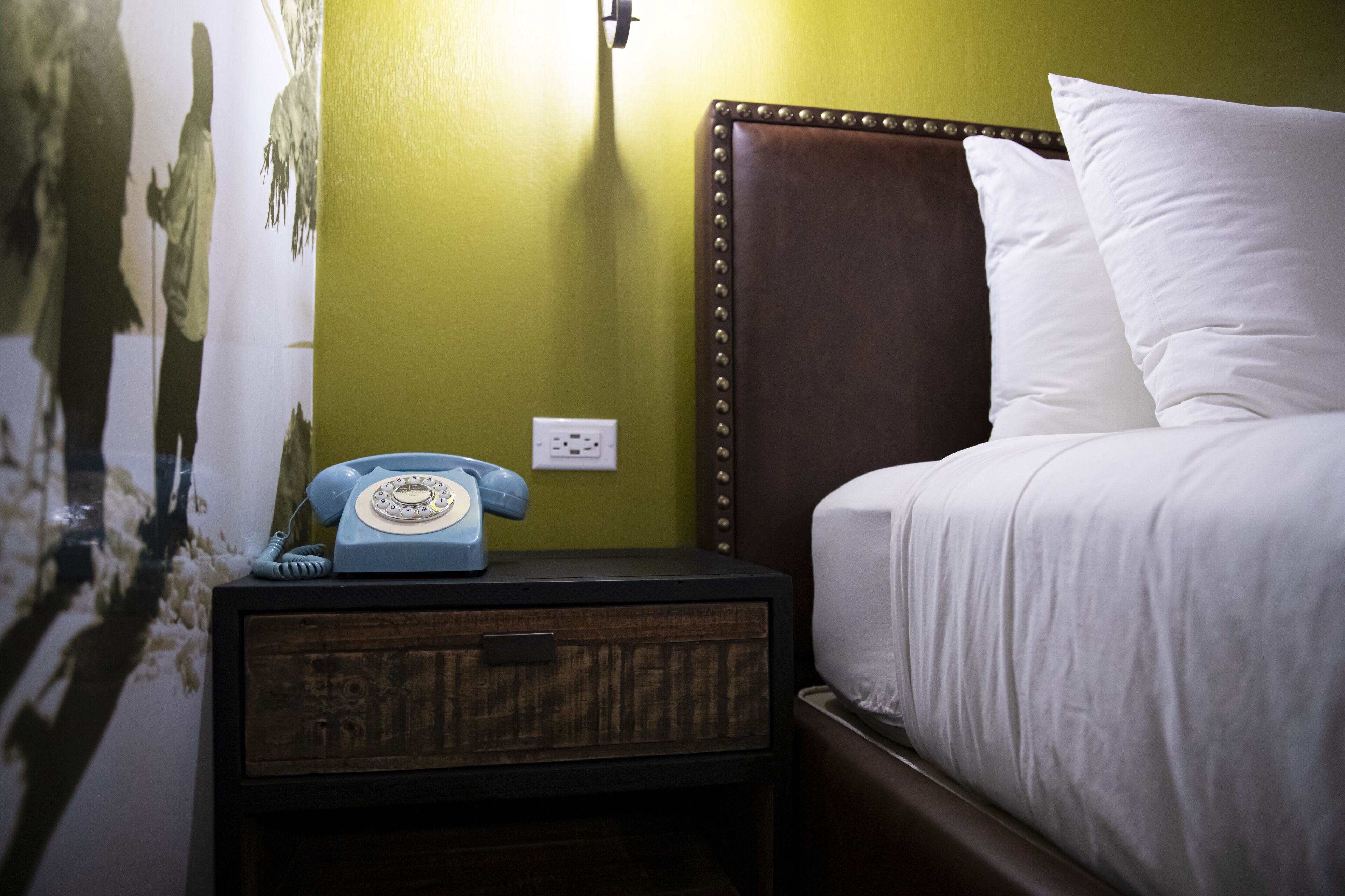 Una cama con sábanas blancas, cabecera marrón y mesita de noche con un teléfono rotativo azul.