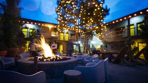 An outdoor seating area with a fire pit, surrounded by chairs and tables, under a tree decorated with lights. - Campfire Hotel (Bend)